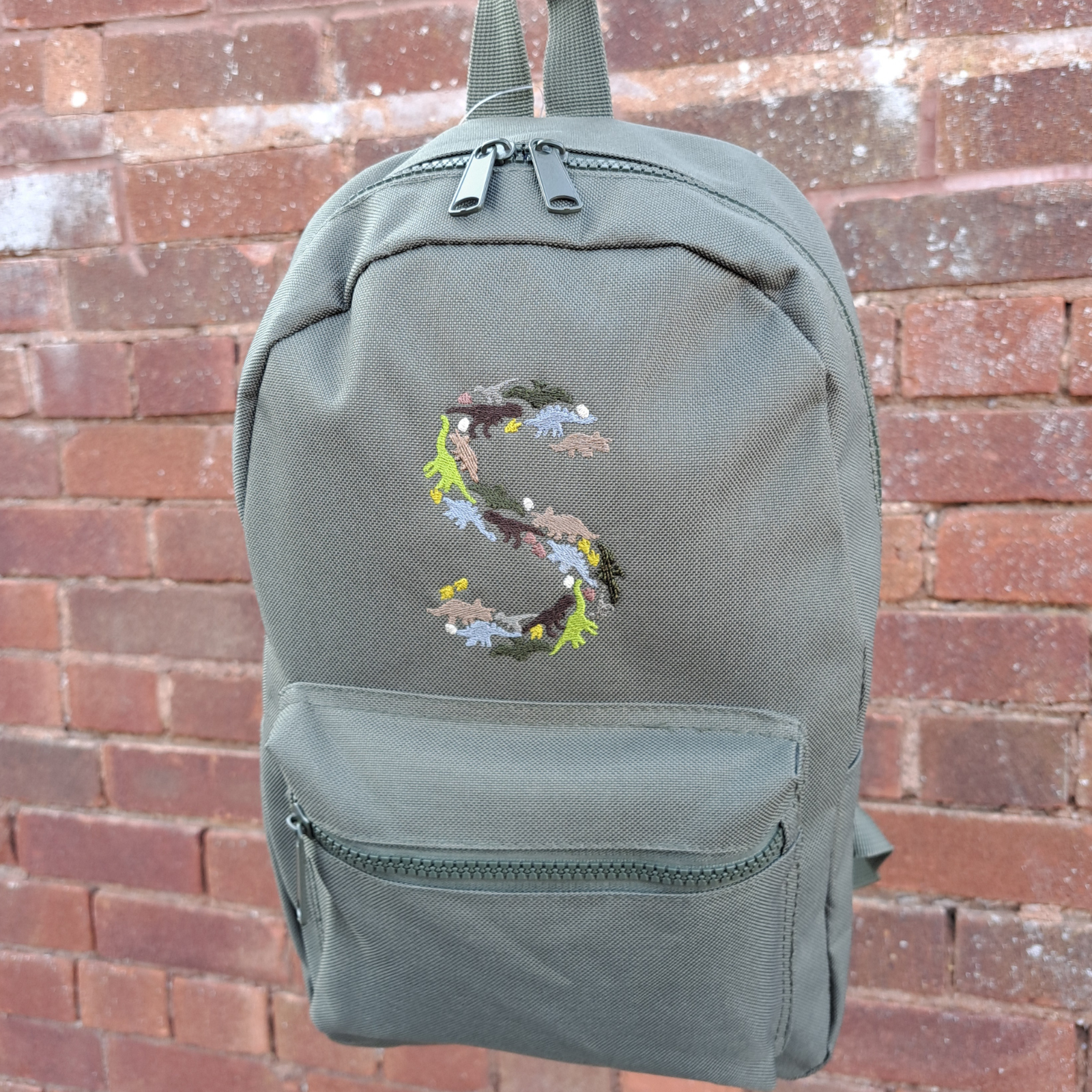 Image of a khaki green child's rucksack which has been embroidered on the front with a capital S which is made up from images of dinosaurs, footprints and eggs, in different coloured threads which compliment the colour of the bag. The bag is available in pistachio, powder pink, powder blue, light grey, fuchsia pink, navy blue, black, lavender and olive green. 