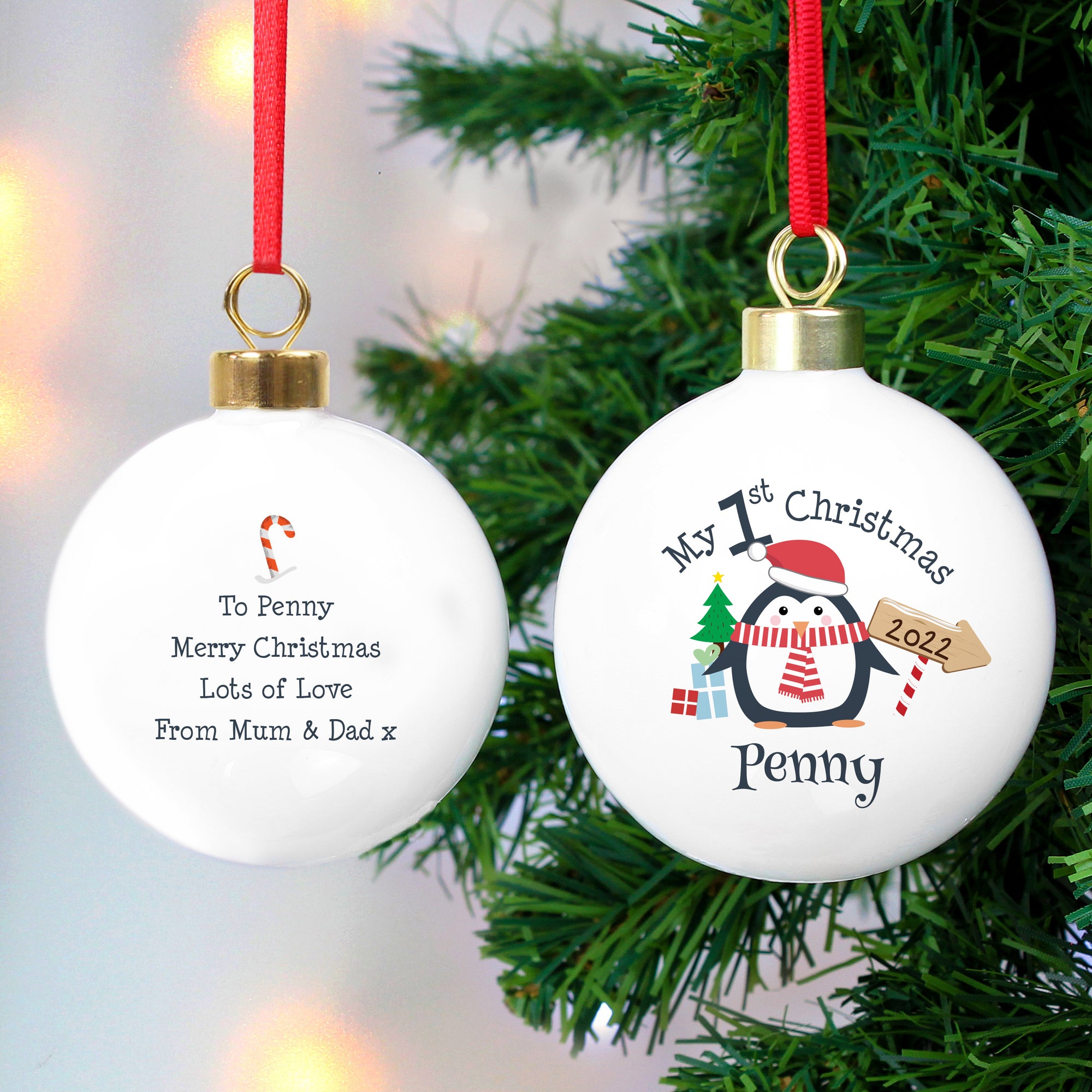 Image of the front and back of a personalised Christmas bauble. The bauble is white, the front features an image of a penguin with a hat on and scarf on. There is a small tree and gifts next to the penguin and an arrow sign with a year. Above the penguin in black text is ‘My 1st Christmas’ and below the penguin you can add a name. The back of the bauble has a small red and white candy cane and below it you can add your own message which will be printed in a black font.