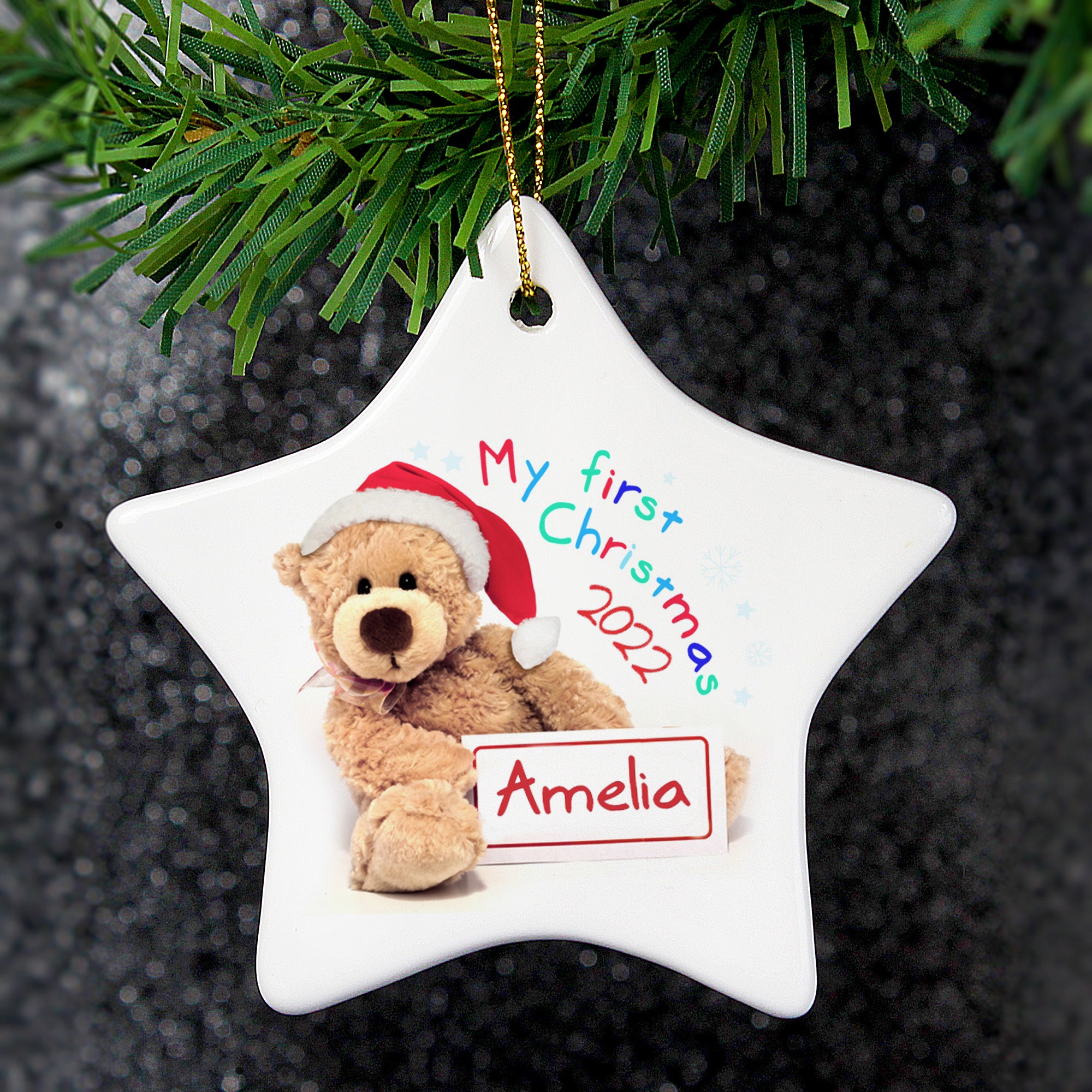 Personalised white ceramic star shaped Christmas decoration with an image of a teddy bear wearing a red and white santa hat and holding a sign which can be personalised with a name of your choice. Next to the bear is the text 'My first Christmas' in bright multi coloured lettering and you can also add a year which will be printed below that in red.