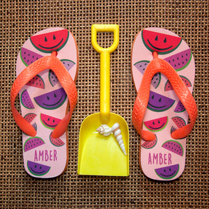 Image of a pair of children's personalised flip flops. The flip flops have an orange sole and straps, and the flip flops have an illustration of bright watermelons in red, pink and purple on a pale pink background. The flip flops can be personalised wtih a name of up to 10 characters on the heel. The flip flops are available in 3 different child sizes.
