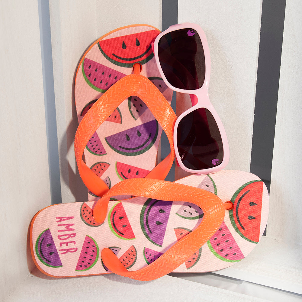 Image of a pair of children's personalised flip flops. The flip flops have an orange sole and straps, and the flip flops have an illustration of bright watermelons in red, pink and purple on a pale pink background. The flip flops can be personalised wtih a name of up to 10 characters on the heel. The flip flops are available in 3 different child sizes.