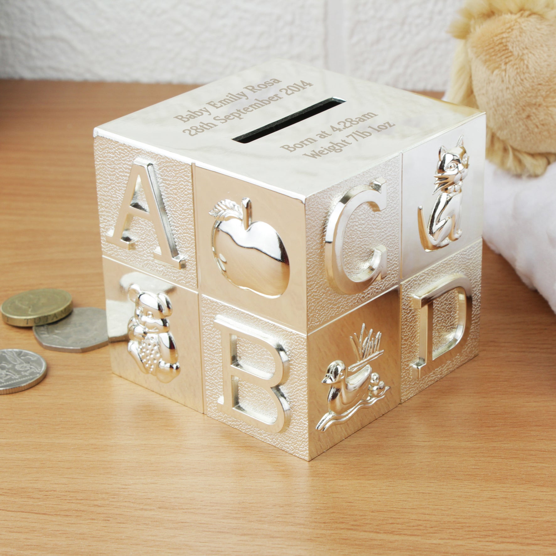A silver-plated engraved square money box. The money box has an ABCD design on the four sides and the top of the money box has the money slot in it. The money box can be engraved with two lines of text above the money slot and two lines of text below the money slot. This moneybox measures 7.7cm by 7.7cm by 7.7cm.
