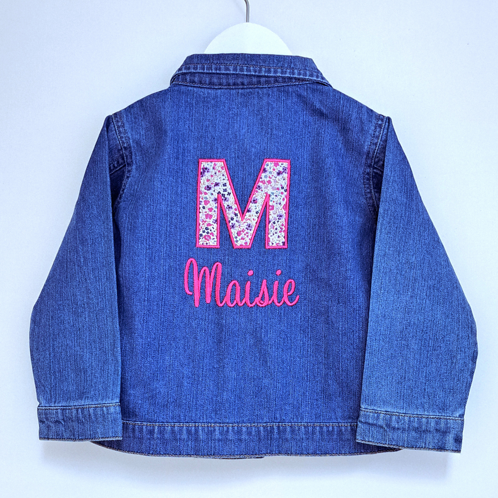 image of a childs denim jacket that has been personalised on the back with an embroidered applique initial using Liberty of London fabric. Below the initial the name has been embroidered in a fuchsia coloured thread.