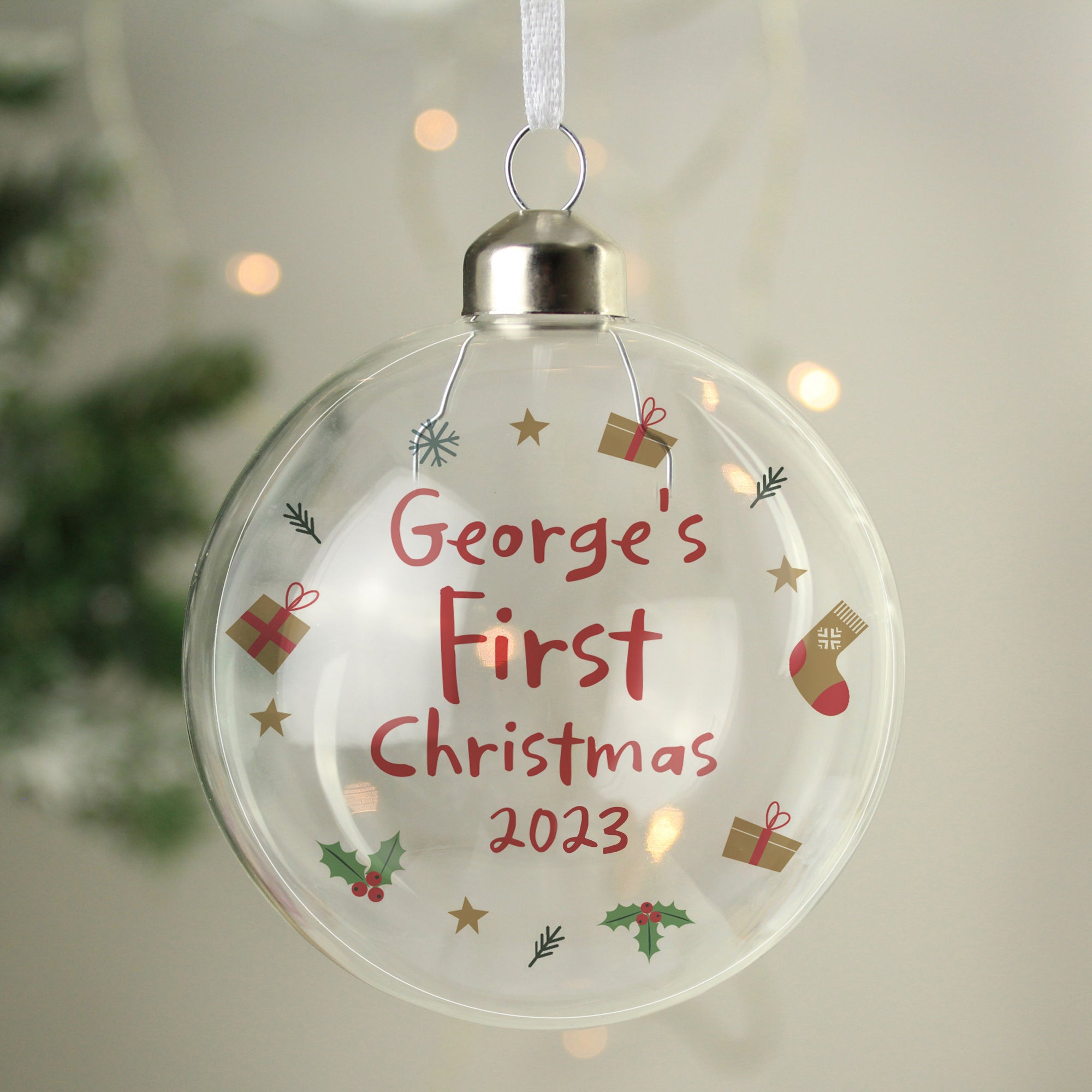 Image of a clear glass Christmas bauble which can be personalised with a name and year of your choice which will be printed in red font in a handwriting style. The bauble features the words 'First Christmas' as a fixed part of the design.