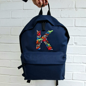 Image of a navy blue rucksack that has been embroidered with a letter K in a dinosaur font. The font is made up of different dinosaurs, footprints and eggs which are embroidered in complementary colours.