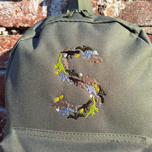 Image of a khaki green child's rucksack which has been embroidered on the front with a capital S which is made up from images of dinosaurs, footprints and eggs, in different coloured threads which compliment the colour of the bag. The bag is available in pistachio, powder pink, powder blue, light grey, fuchsia pink, navy blue, black, lavender and olive green.