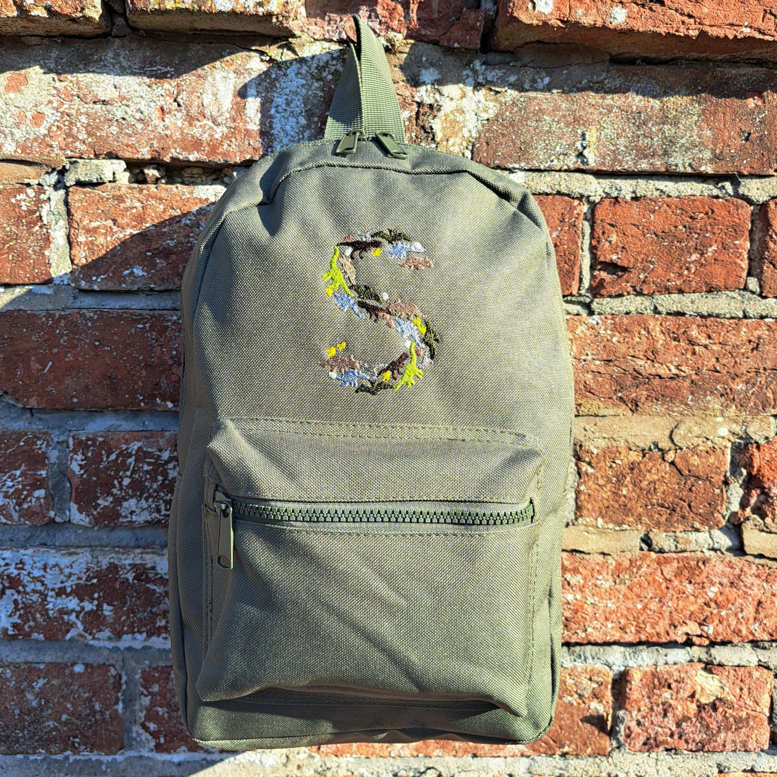 Image of a khaki green child's rucksack which has been embroidered on the front with a capital S which is made up from images of dinosaurs, footprints and eggs, in different coloured threads which compliment the colour of the bag. The bag is available in pistachio, powder pink, powder blue, light grey, fuchsia pink, navy blue, black, lavender and olive green.