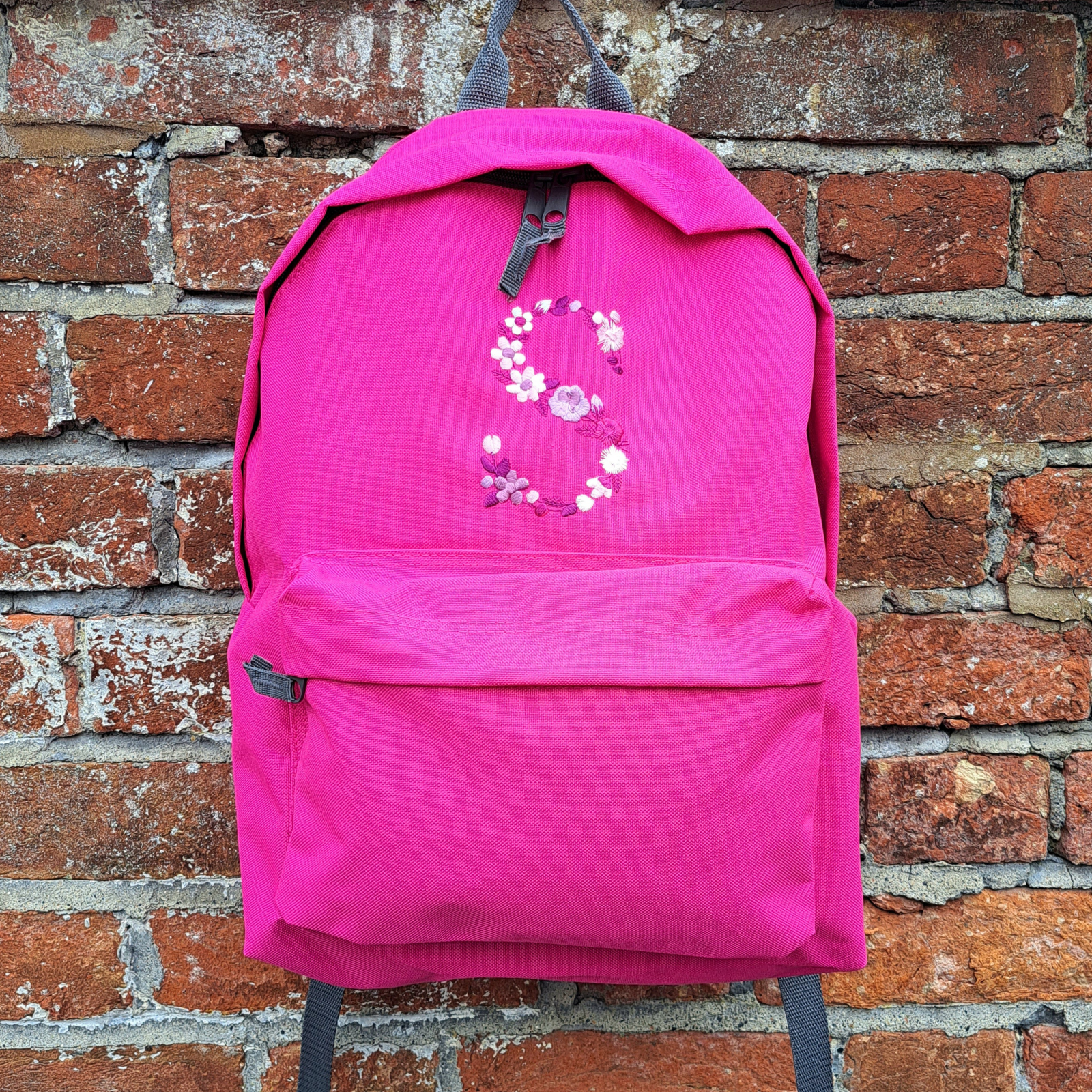 Image of a fuchsia pink coloured junior rucksack that has been embroidered with a capital S which is formed from many different smaller flowers and leaves.