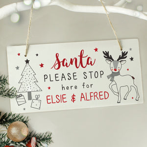 Image of a white wooden Christmas Eve plaque that is printed with the text 'Santa please stop here for' which is fixed and you can then add your own names. The sign features a modern image of a Chrismtas tree with gifts underneath it as well as a reindeer.