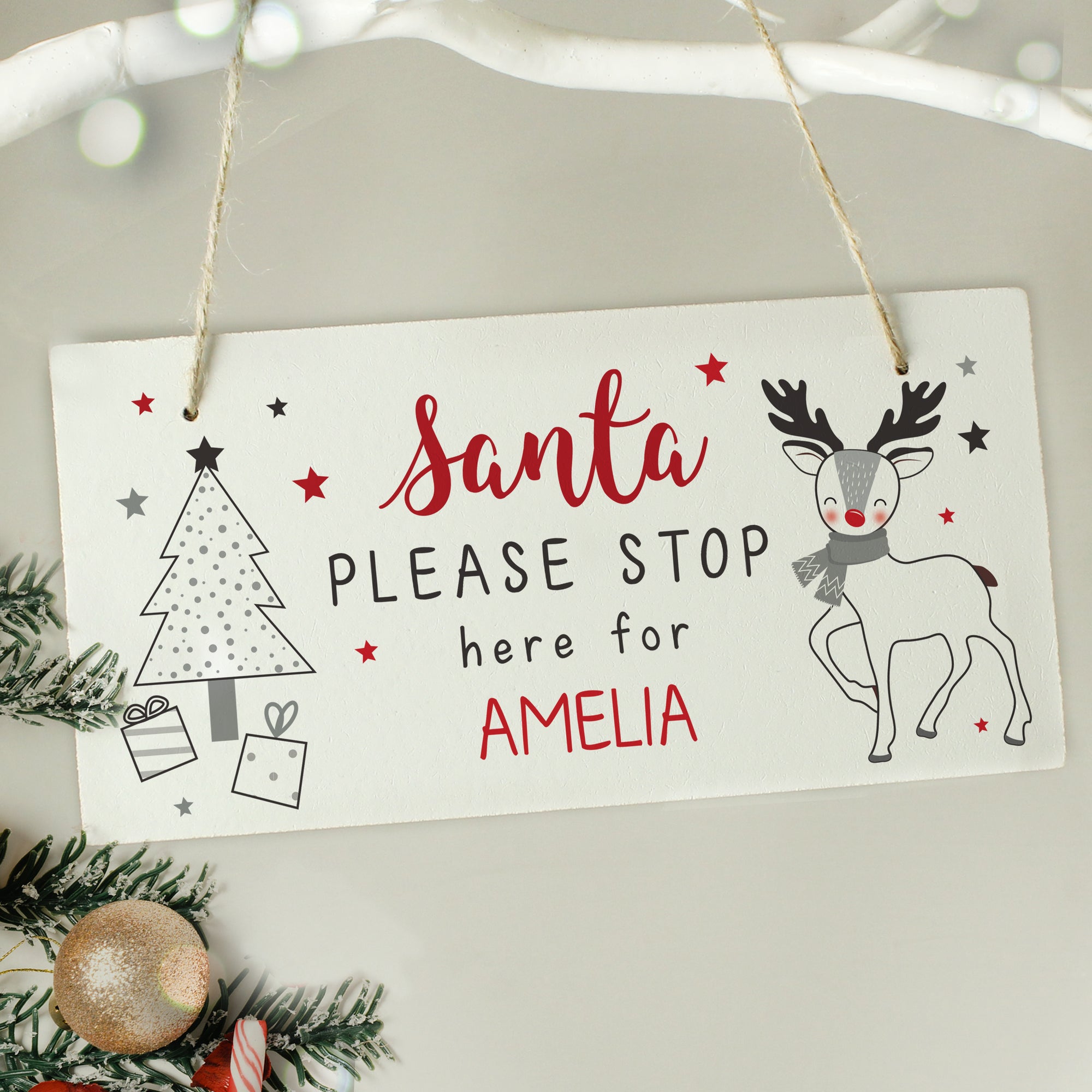 Image of a white wooden Christmas Eve plaque that is printed with the text 'Santa please stop here for' which is fixed and you can then add your own names. The sign features a modern image of a Chrismtas tree with gifts underneath it as well as a reindeer.