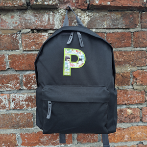 Image of a black school backpack that has been personalised on the front with an initial P in Liberty fabric which is appliqued onto the back with green coordinating embroidery thread. The bag has a zipped main compartment and a smaller zipped pocket on the front along with two adjustable back straps.