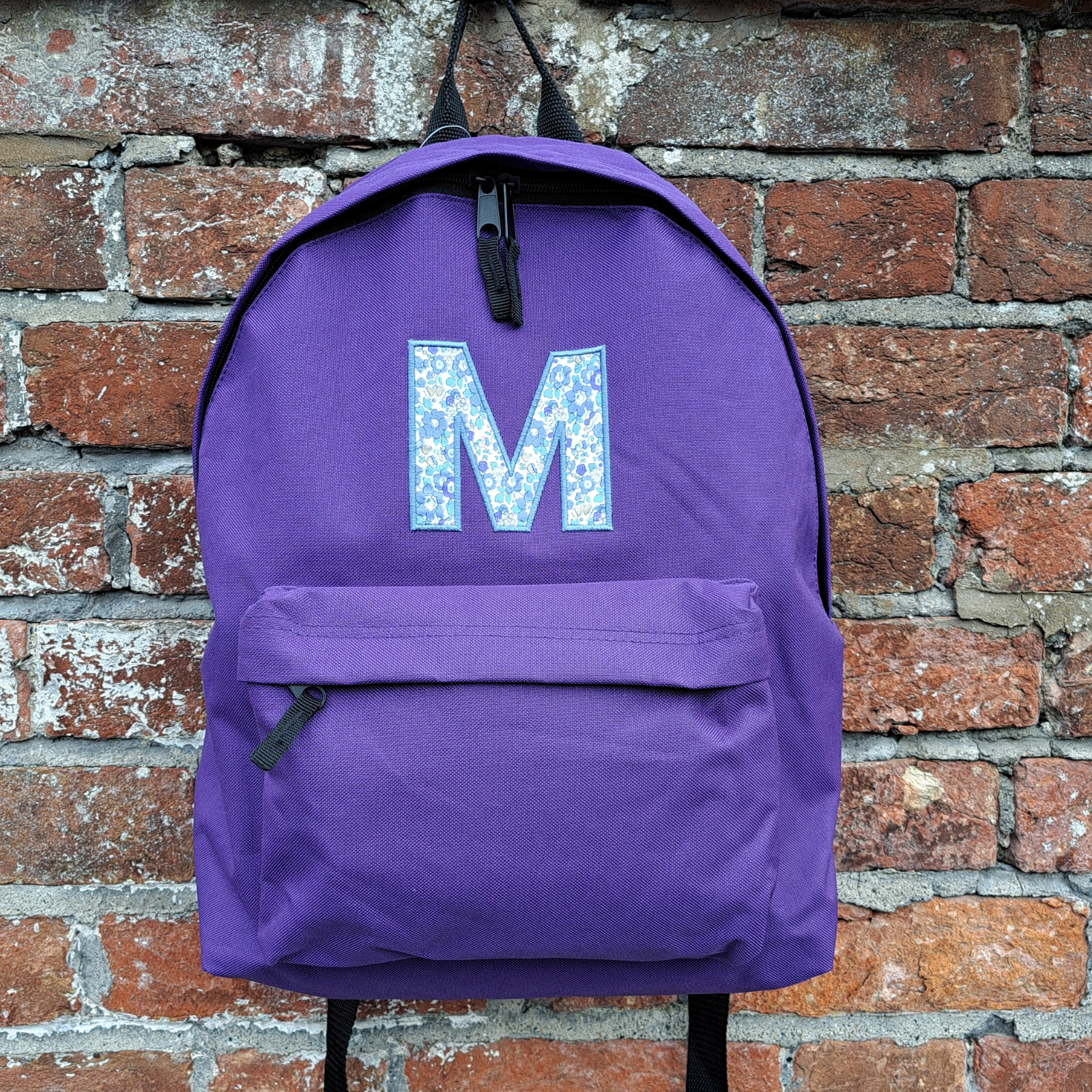 Image of a purple school backpack that has been personalised on the front with an initial M in Liberty of London fabric which is appliqued onto the bag with blue coordinating embroidery thread. The bag has a zipped main compartment and a smaller zipped pocket on the front along with two adjustable back straps.