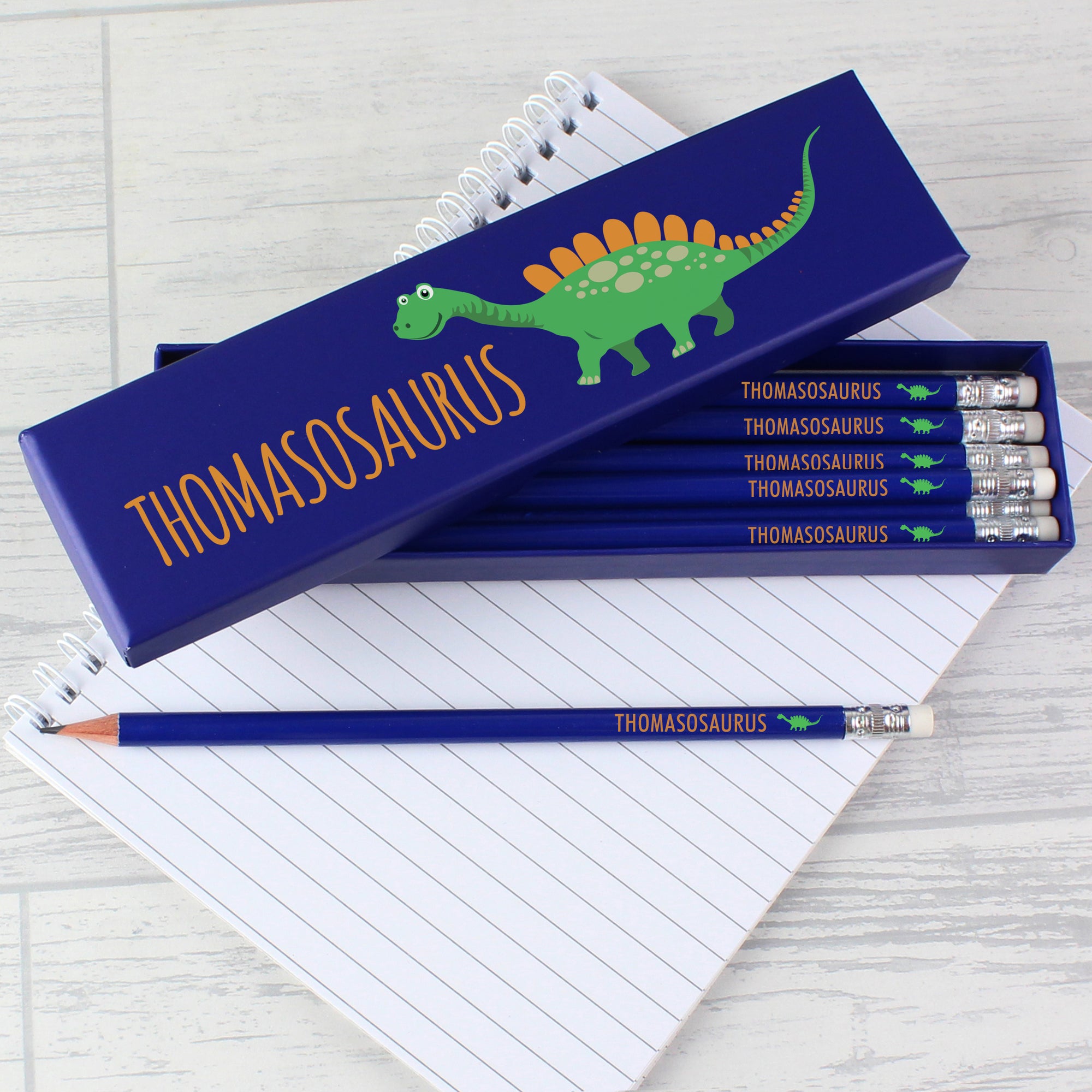Image of a blue box with a friendly dinosaur image on the front and a name of your choice which will be printed in orange uppercase letters. Inside the box there are 12 blue HB writing pencils, each with a rubber on the end which can also be personalised with a name of your choice and printed in orange uppercase letters which will be followed by a mini dinosaur image.