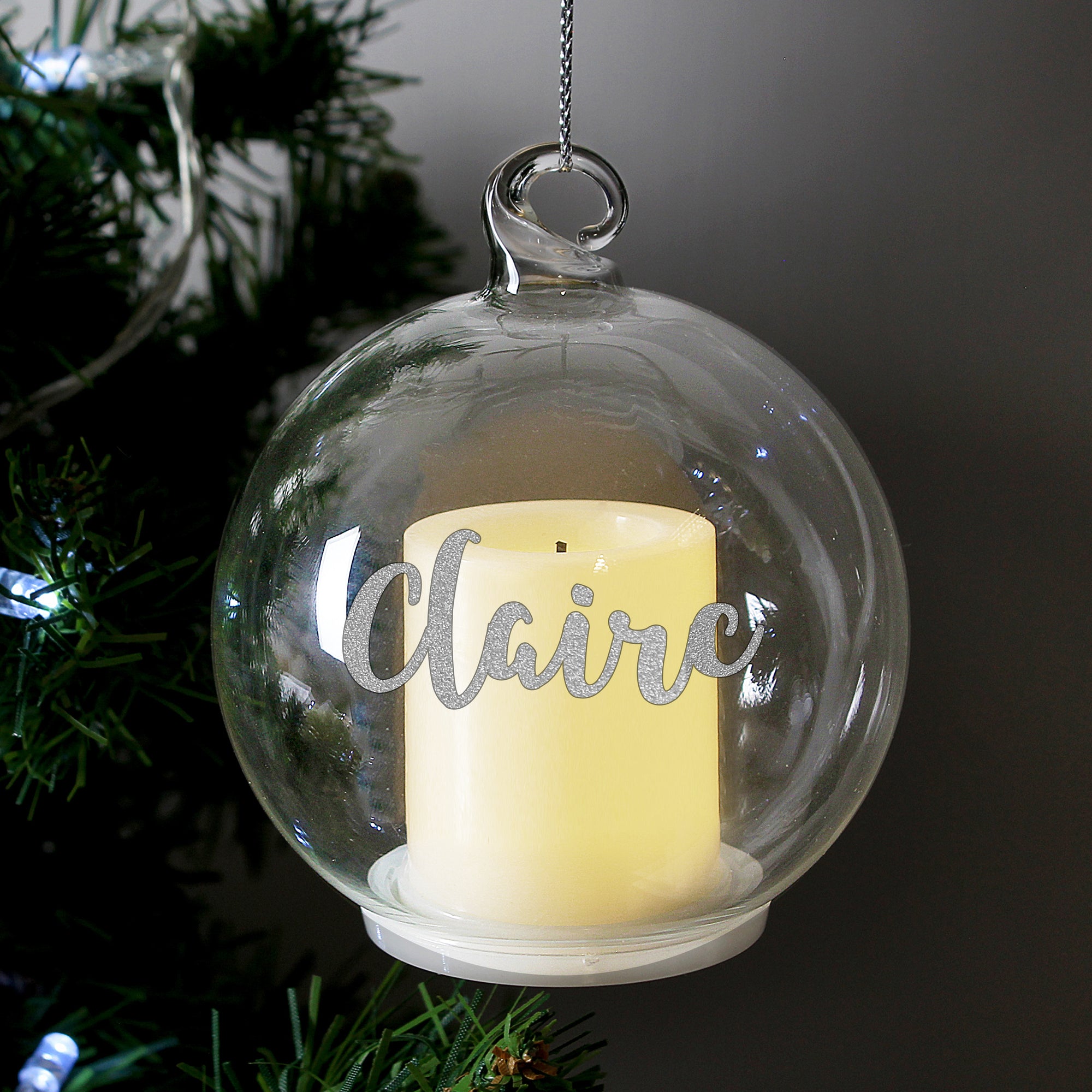 Personalised clear glass round Christmas bauble which has a battery operated cream yellow LED candle inside.  The bauble can be personalsied with a name in a silver hand-written styled modern font and comes with a silver thread to hang it up.