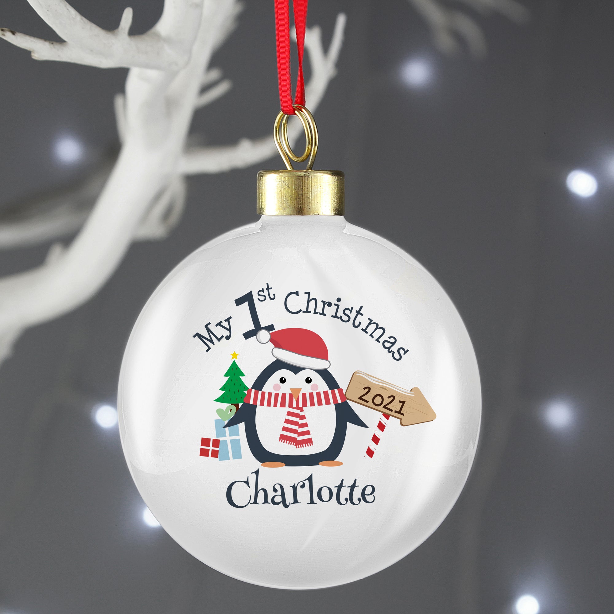 Image of the front of a personalised Christmas bauble. The bauble is white with a gold top. The front of the bauble features an image of a penguin with a Santa hat on and a scarf. There is a little Christmas tree and gifts next to the penguin and an arrow sign which can be personalised with a year. Above the penguin in black text is ‘My 1st Christmas’ and below the penguin you can add a name. 