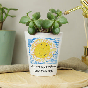 Image of a white ceramic plant pot with a plant inside it. The plant pot can be printed with your child's own artwork that will be printed on the front and there is space underneath the artwork for you own optional text which will be printed in a black handwritten style font.