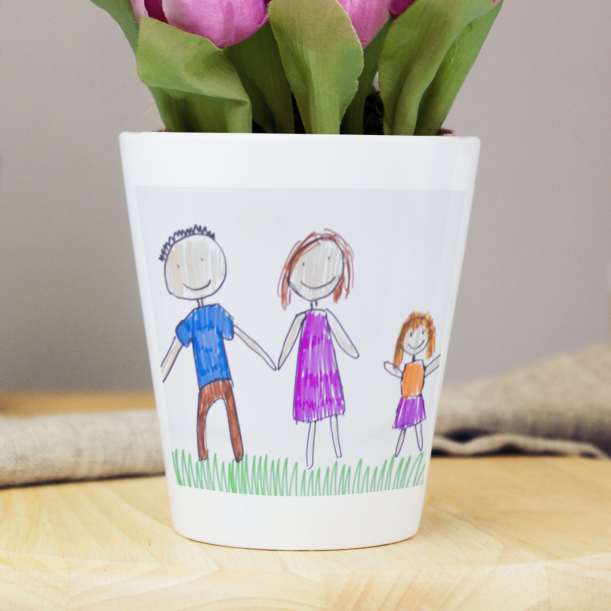 Image of a white ceramic plant pot with a plant inside it. The plant pot can be printed with your child's own artwork that will be printed on the front and there is space underneath the artwork for you own optional text which will be printed in a black handwritten style font.