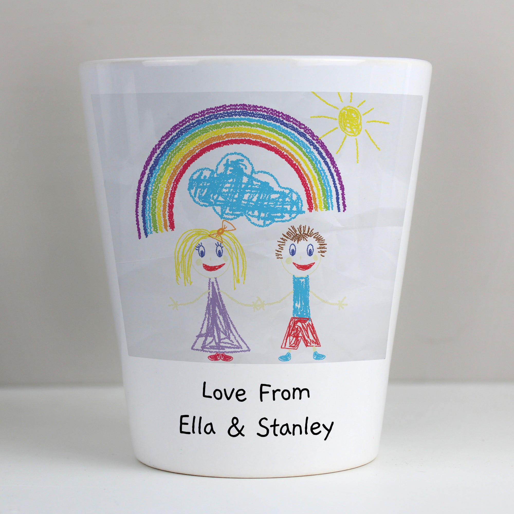 Image of a white ceramic plant pot with a plant inside it. The plant pot can be printed with your child's own artwork that will be printed on the front and there is space underneath the artwork for you own optional text which will be printed in a black handwritten style font.