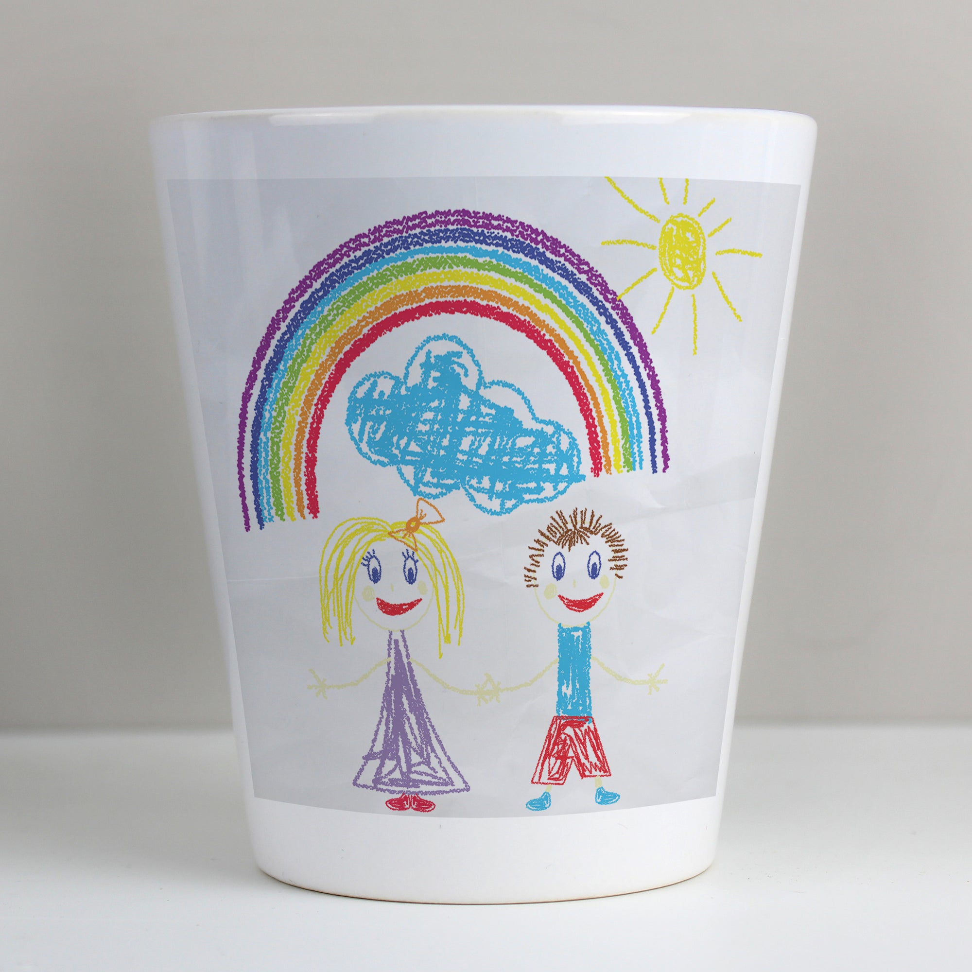 Image of a white ceramic plant pot with a plant inside it. The plant pot can be printed with your child's own artwork that will be printed on the front and there is space underneath the artwork for you own optional text which will be printed in a black handwritten style font.