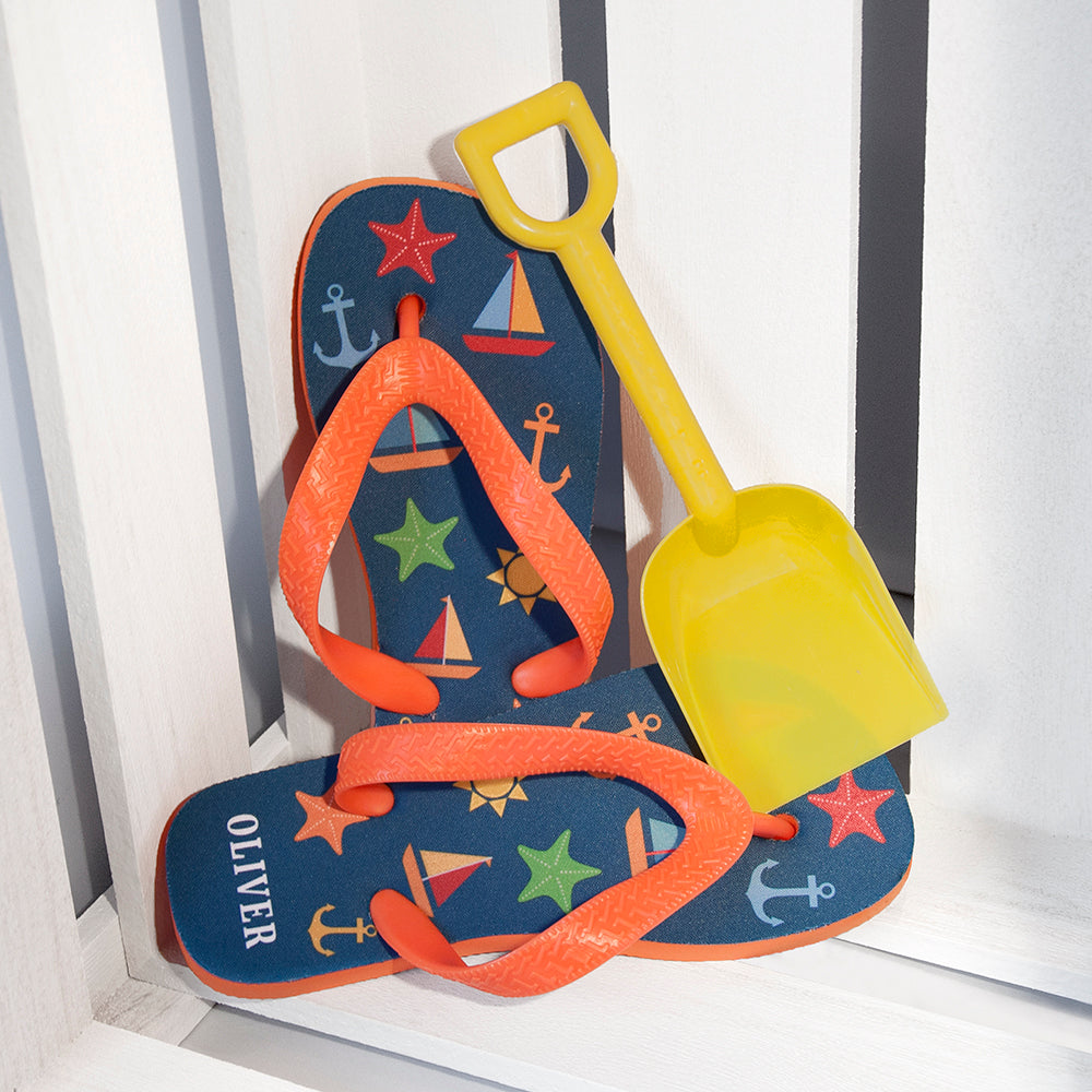 Image of a pair of children's personalised flip flops. The flip flops have an orange sole and straps, and the flip flops have illustrations of bright beach themed items such as sail boats, star fish, anchors and a sun on a blue background. The flip flops can be personalised wtih a name of up to 10 characters on the heel. The flip flops are available in 3 different child sizes.