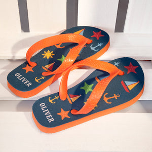 Image of a pair of children's personalised flip flops. The flip flops have an orange sole and straps, and the flip flops have illustrations of bright beach themed items such as sail boats, star fish, anchors and a sun on a blue background. The flip flops can be personalised wtih a name of up to 10 characters on the heel. The flip flops are available in 3 different child sizes.