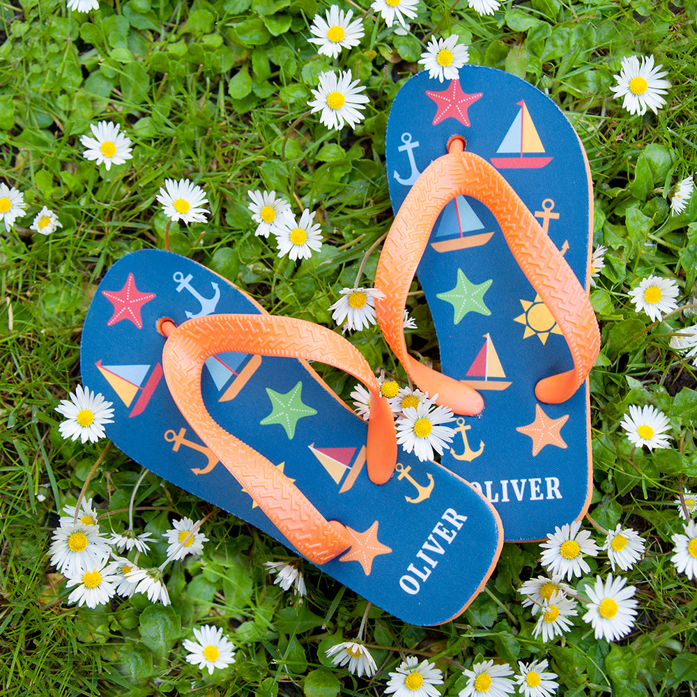 Image of a pair of children's personalised flip flops. The flip flops have an orange sole and straps, and the flip flops have illustrations of bright beach themed items such as sail boats, star fish, anchors and a sun on a blue background. The flip flops can be personalised wtih a name of up to 10 characters on the heel. The flip flops are available in 3 different child sizes.