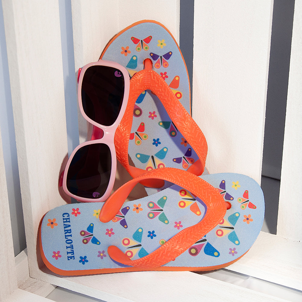 Image of a pair of children's personalised flip flops. The flip flops have an orange sole and straps, and the flip flops have an illustration of bright butterflies on a pale blue background. The flip flops can be personalised wtih a name of up to 10 characters on the heel. The flip flops are available in 3 different child sizes.