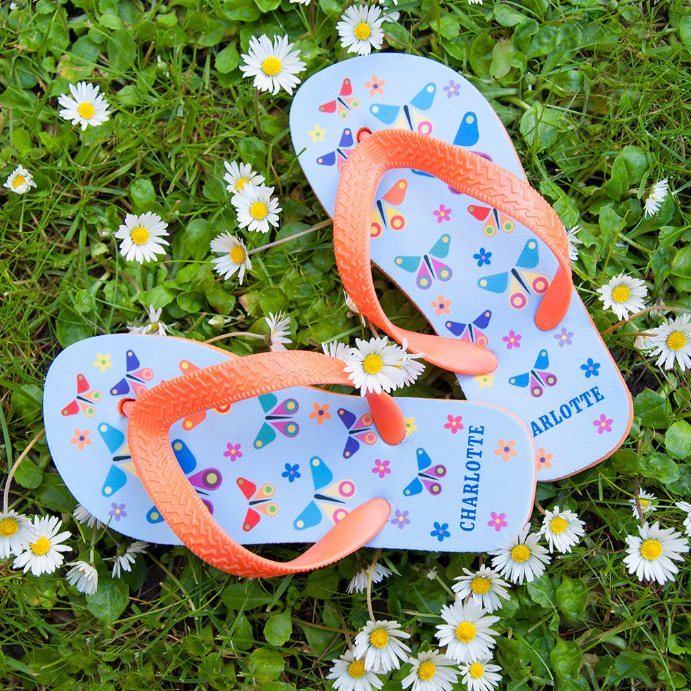 Image of a pair of children's personalised flip flops. The flip flops have an orange sole and straps, and the flip flops have an illustration of bright butterflies on a pale blue background. The flip flops can be personalised wtih a name of up to 10 characters on the heel. The flip flops are available in 3 different child sizes.