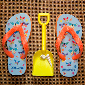 Image of a pair of children's personalised flip flops. The flip flops have an orange sole and straps, and the flip flops have an illustration of bright butterflies on a pale blue background. The flip flops can be personalised wtih a name of up to 10 characters on the heel. The flip flops are available in 3 different child sizes.