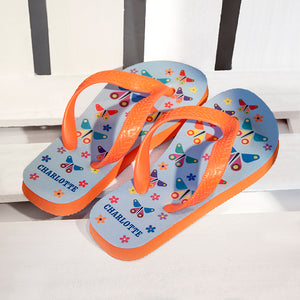 Image of a pair of children's personalised flip flops. The flip flops have an orange sole and straps, and the flip flops have an illustration of bright butterflies on a pale blue background. The flip flops can be personalised wtih a name of up to 10 characters on the heel. The flip flops are available in 3 different child sizes.
