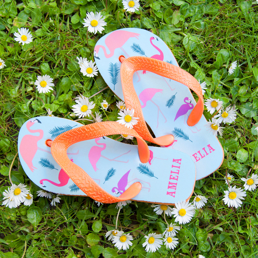 Image of a pair of children's personalised flip flops. The flip flops have an orange sole and straps, and the flip flops have an illustration of bright flamingos on a pale blue background. The flip flops can be personalised wtih a name of up to 10 characters on the heel. The flip flops are available in 3 different child sizes.