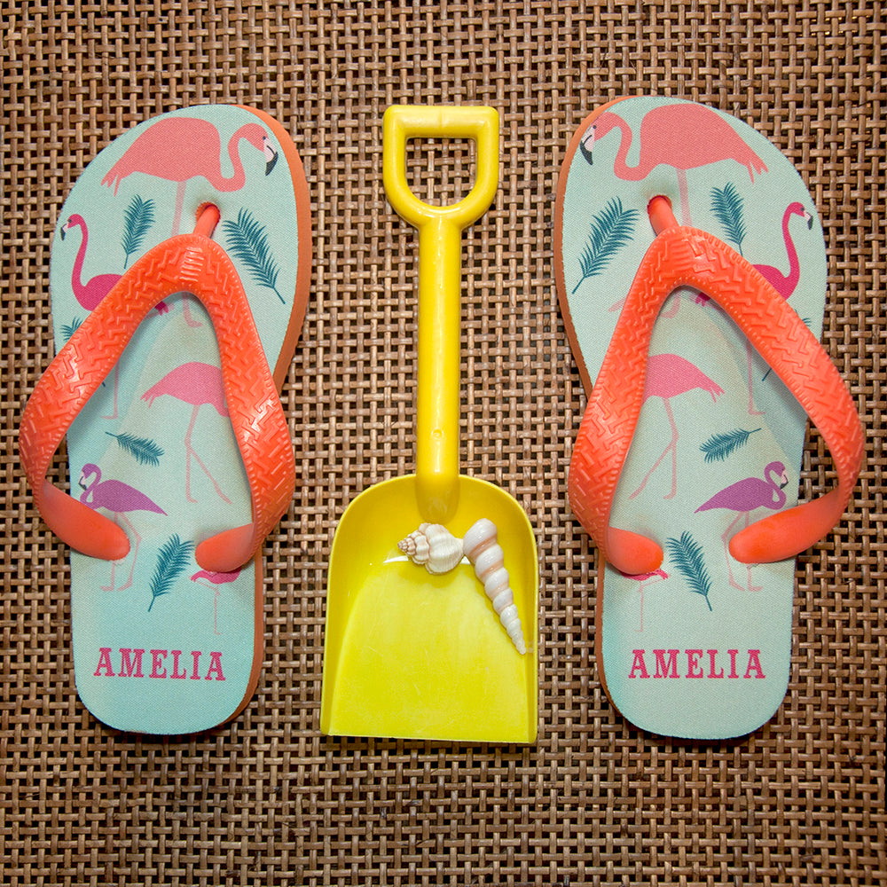Image of a pair of children's personalised flip flops. The flip flops have an orange sole and straps, and the flip flops have an illustration of bright flamingos on a pale blue background. The flip flops can be personalised wtih a name of up to 10 characters on the heel. The flip flops are available in 3 different child sizes.