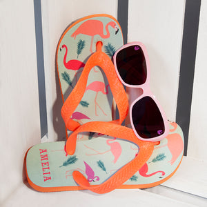 Image of a pair of children's personalised flip flops. The flip flops have an orange sole and straps, and the flip flops have an illustration of bright flamingos on a pale blue background. The flip flops can be personalised wtih a name of up to 10 characters on the heel. The flip flops are available in 3 different child sizes.