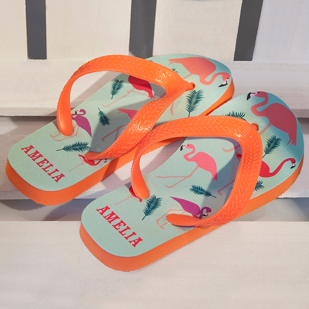 Image of a pair of children's personalised flip flops. The flip flops have an orange sole and straps, and the flip flops have an illustration of bright flamingos on a pale blue background. The flip flops can be personalised wtih a name of up to 10 characters on the heel. The flip flops are available in 3 different child sizes.