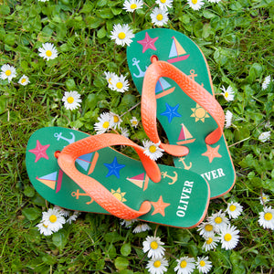 Image of a pair of children's personalised flip flops. The flip flops have an orange sole and straps, and the flip flops have illustrations of bright beach themed items such as sail boats, star fish, anchors and a sun on a green background. The flip flops can be personalised wtih a name of up to 10 characters on the heel. The flip flops are available in 3 different child sizes.