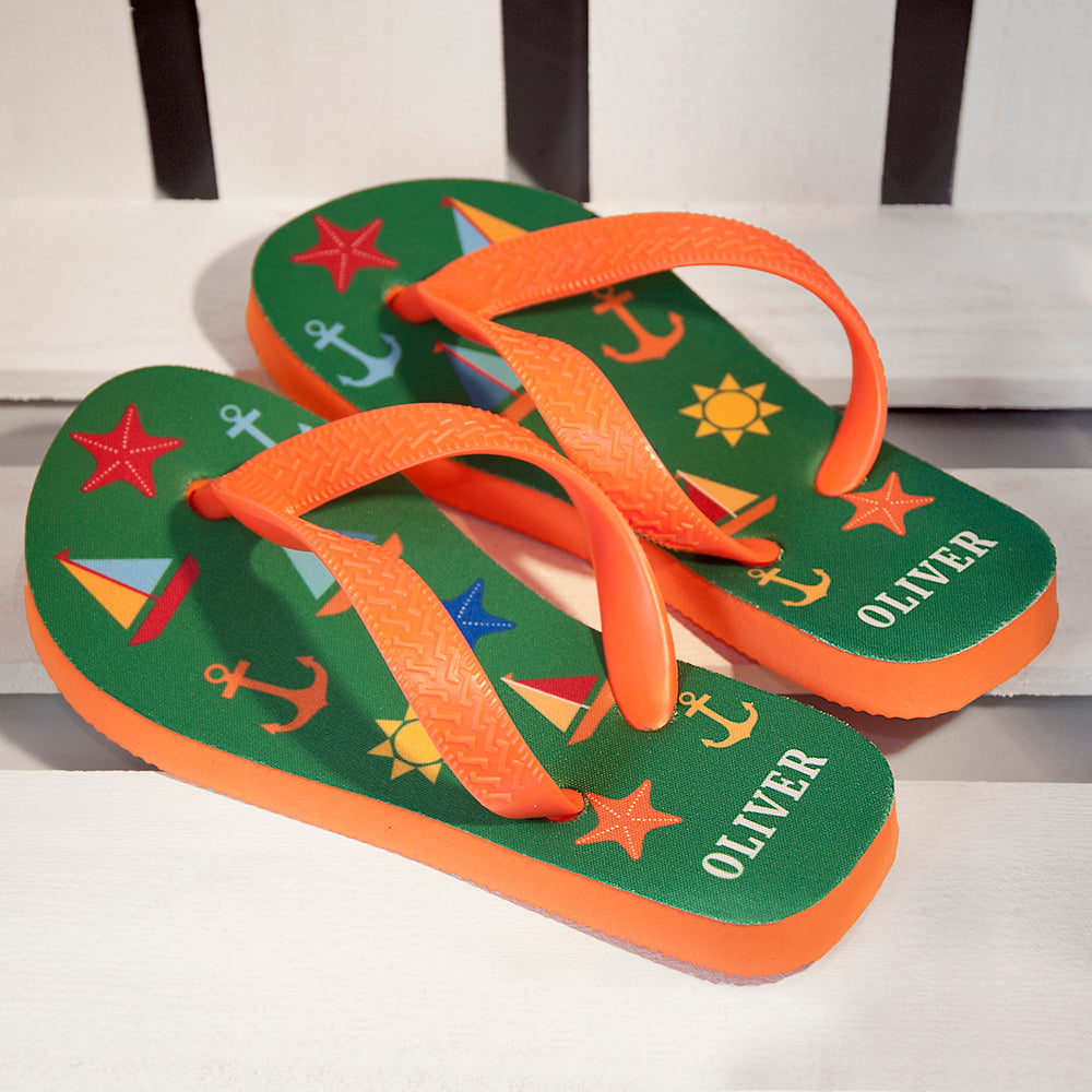 Image of a pair of children's personalised flip flops. The flip flops have an orange sole and straps, and the flip flops have illustrations of bright beach themed items such as sail boats, star fish, anchors and a sun on a green background. The flip flops can be personalised wtih a name of up to 10 characters on the heel. The flip flops are available in 3 different child sizes.
