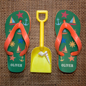 Image of a pair of children's personalised flip flops. The flip flops have an orange sole and straps, and the flip flops have illustrations of bright beach themed items such as sail boats, star fish, anchors and a sun on a green background. The flip flops can be personalised wtih a name of up to 10 characters on the heel. The flip flops are available in 3 different child sizes.