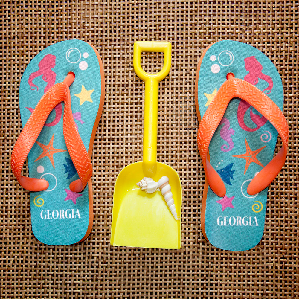 Image of a pair of children's personalised flip flops. The flip flops have an orange sole and straps, and the flip flops have an illustration of bright fish, star fish and a mermaid on a pale blue background. The flip flops can be personalised wtih a name of up to 10 characters on the heel. The flip flops are available in 3 different child sizes.