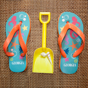 Image of a pair of children's personalised flip flops. The flip flops have an orange sole and straps, and the flip flops have an illustration of bright fish, star fish and a mermaid on a pale blue background. The flip flops can be personalised wtih a name of up to 10 characters on the heel. The flip flops are available in 3 different child sizes.