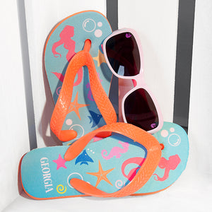 Image of a pair of children's personalised flip flops. The flip flops have an orange sole and straps, and the flip flops have an illustration of bright fish, star fish and a mermaid on a pale blue background. The flip flops can be personalised wtih a name of up to 10 characters on the heel. The flip flops are available in 3 different child sizes.