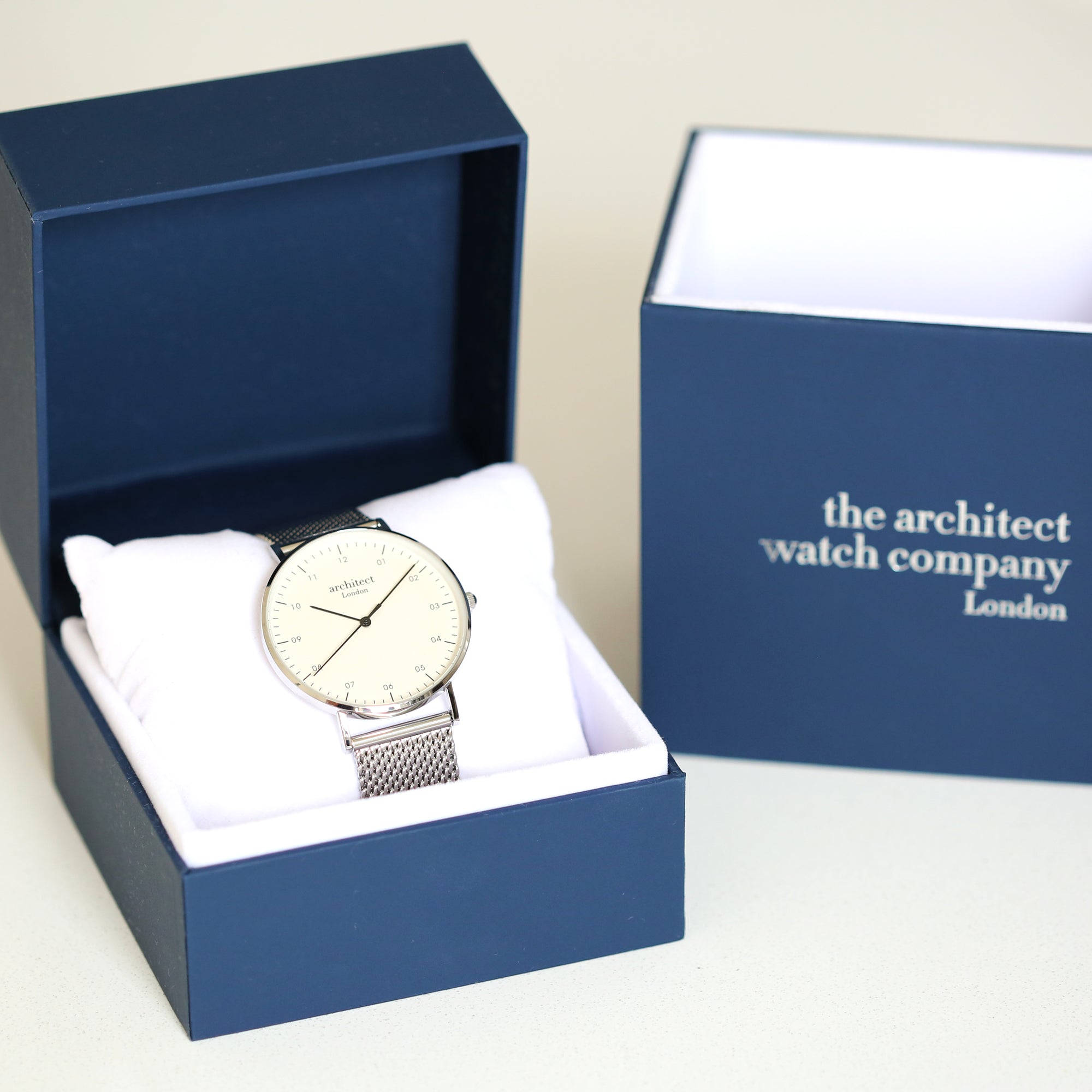 Image of a man's watch in a blue presentation box. The watch can be engraved on the back with your own handwriting. The watch is made by The Architect Watch Company London and has a large white face and a stainless steel mesh strap. The rear of the watch can be engraved with your message written in your own handwriting.