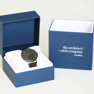 Image of a man's watch in a blue presentation box. The watch can be engraved on the back with your own handwriting. The watch is made by The Architect Watch Company London and has a large black face and a black stainless steel mesh strap. The rear of the watch can be engraved with your message written in your own handwriting.