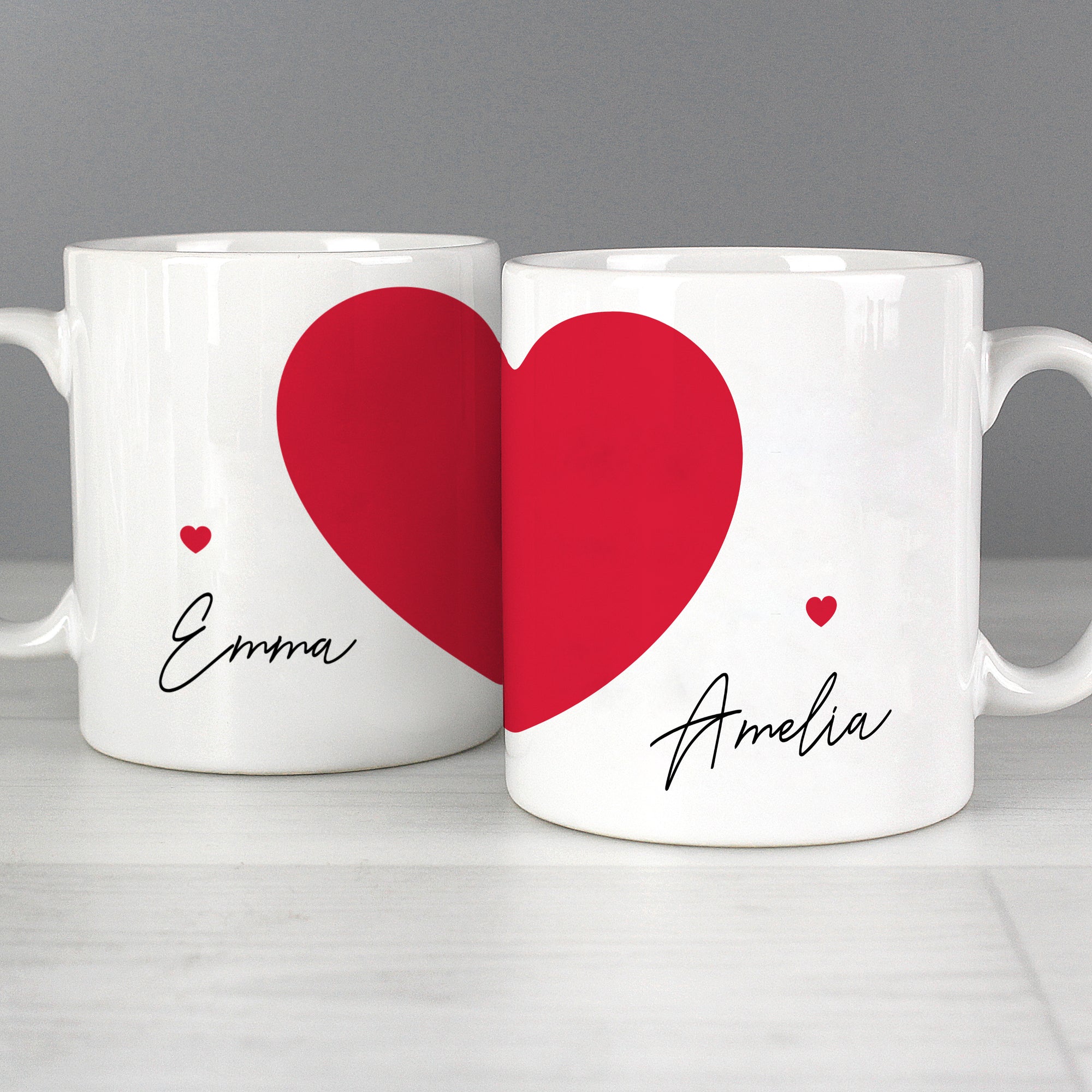 Image of a pair of white ceramic mugs and each mug has a large red heart printed on it as well as a smaller red heart.  Each mug can be personalised with a name of your choice which will be printed in a black handwriting font.