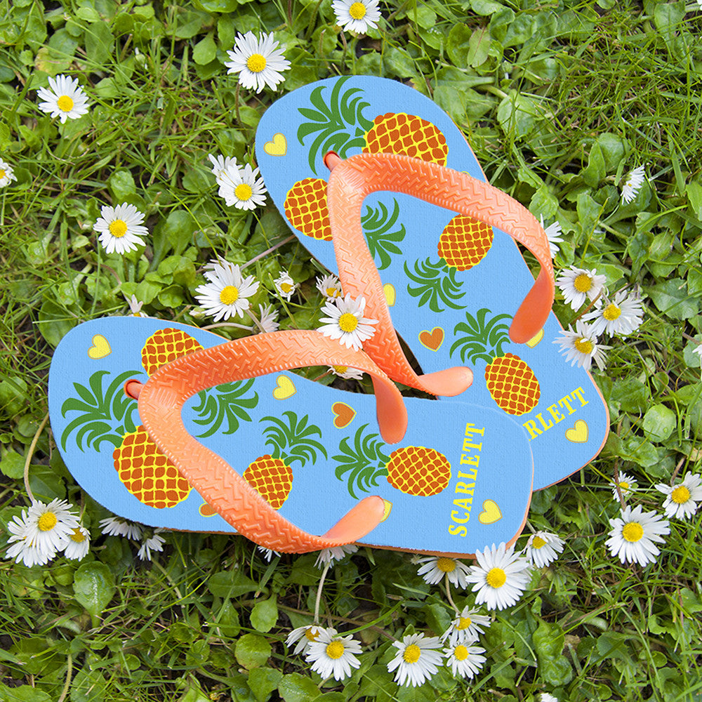 Image of a pair of children's personalised flip flops. The flip flops have an orange sole and straps, and the flip flops have an illustration of bright pineapples and love hearts on a pale blue background. The flip flops can be personalised wtih a name of up to 10 characters on the heel. The flip flops are available in 3 different child sizes.