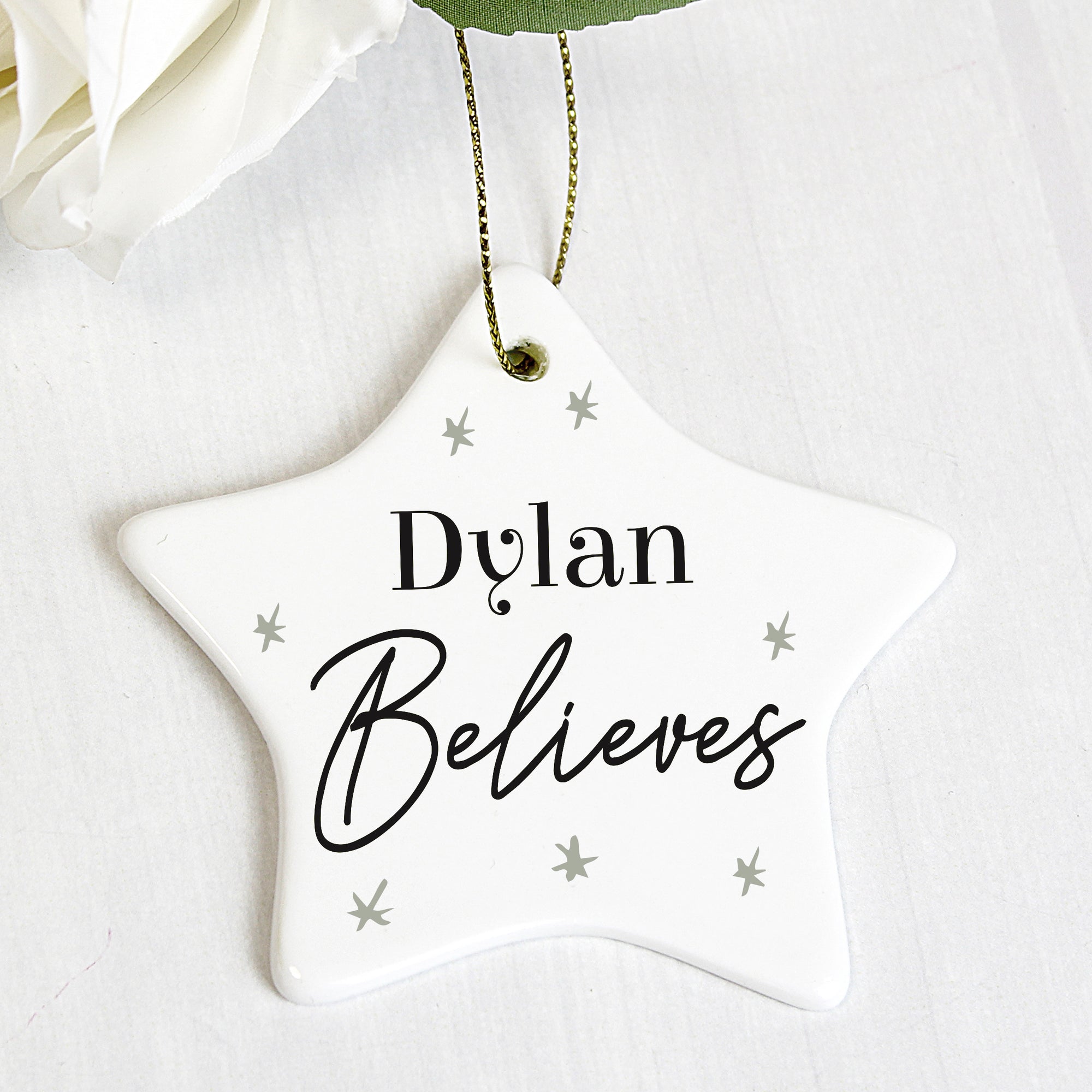 White ceramic star shaped Christmas decoration that comes with a string ready to hang. The decoration can be personalised with a name of up to 12 characters on the front followed by the word 'Believes'. The star has a small number of grey stars printed ono the front in a minimalistic hand drawn style.