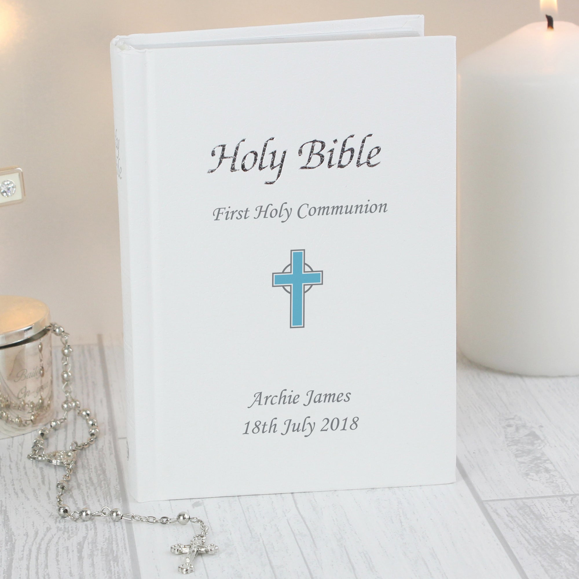 Image of a holy bible with a white leatherette cover featuring the text 'Holy Bible' and a blue cross. The cover can be personalised with 3 lines of text, one line will appear above the image of the cross and the remaining two lines will appear below the image of the cross. The text is printed in silver and the bible has a ribbon bookmark and the pages are edged in silver leaf.