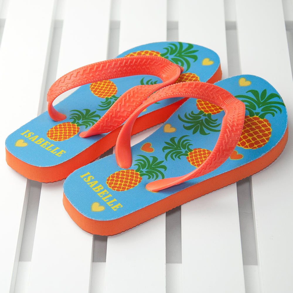 Image of a pair of children's personalised flip flops. The flip flops have an orange sole and straps, and the flip flops have an illustration of bright pineapples and love hearts on a pale blue background. The flip flops can be personalised wtih a name of up to 10 characters on the heel. The flip flops are available in 3 different child sizes.