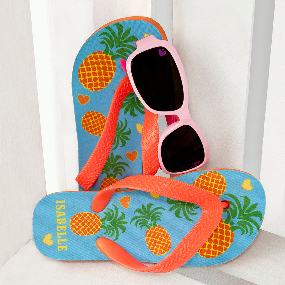 Image of a pair of children's personalised flip flops. The flip flops have an orange sole and straps, and the flip flops have an illustration of bright pineapples and love hearts on a pale blue background. The flip flops can be personalised wtih a name of up to 10 characters on the heel. The flip flops are available in 3 different child sizes.