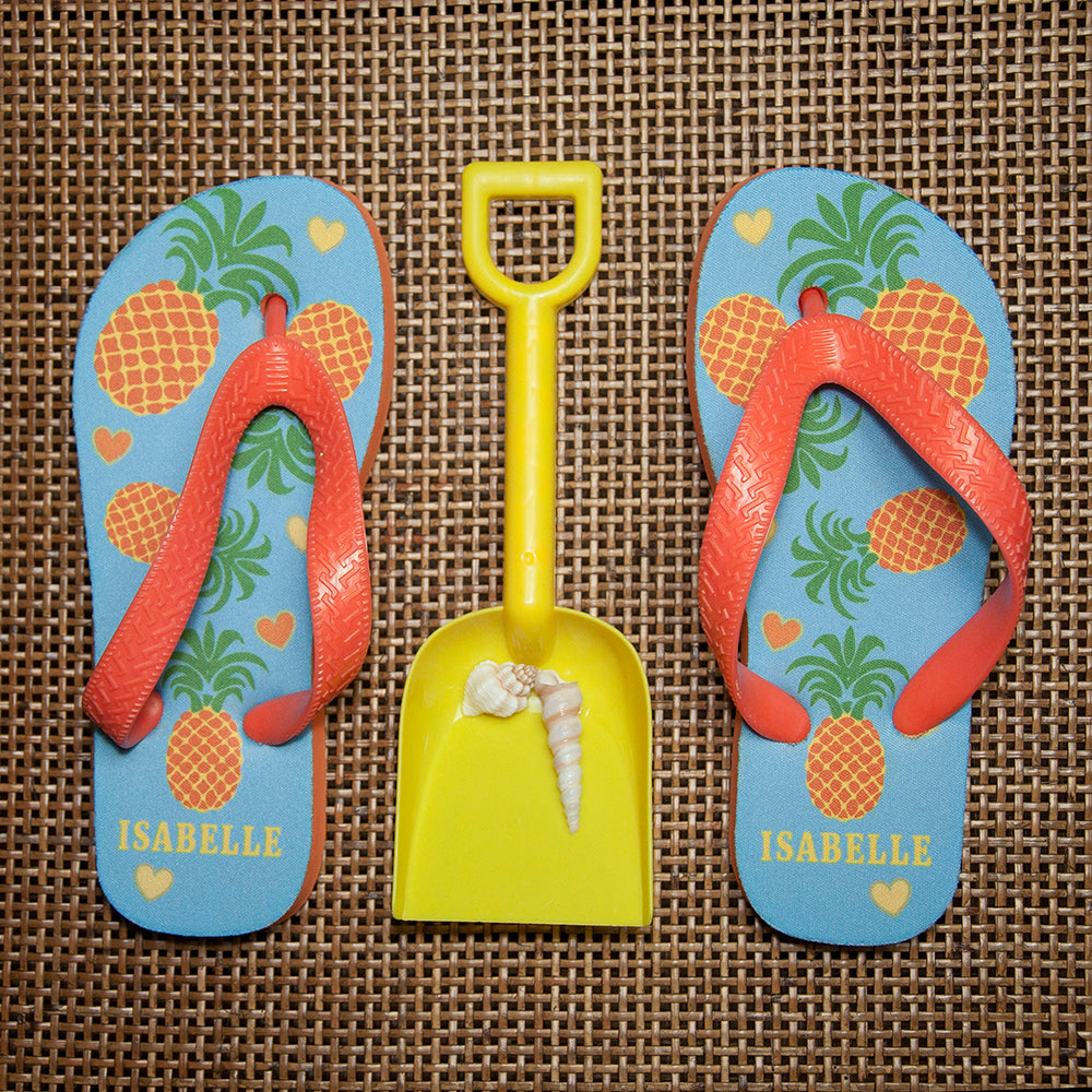 Image of a pair of children's personalised flip flops. The flip flops have an orange sole and straps, and the flip flops have an illustration of bright pineapples and love hearts on a pale blue background. The flip flops can be personalised wtih a name of up to 10 characters on the heel. The flip flops are available in 3 different child sizes.