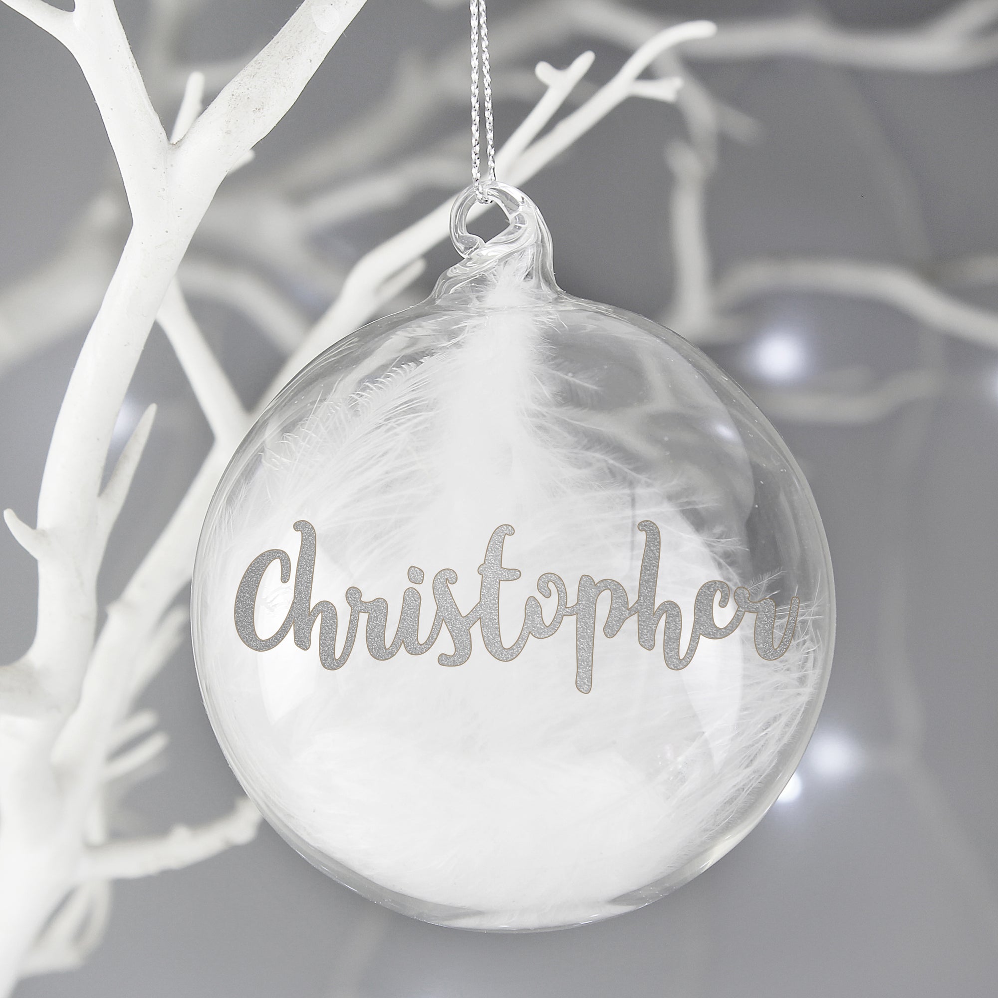 Personalised hand-made clear glass Christmas bauble that has a white feather inside it. The front of the bauble can be personalised with a name of your choice of up to 11 characters that will be printed in a silver glittery modern cursive font. The bauble comes with a string ready to hang and measures approximately 8 cm wide.